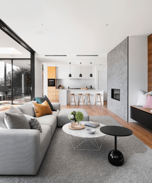 Nowoczesny salon z jasnymi ścianami, szarą sofą, kolorowymi poduszkami i stolikiem kawowym, w tle otwarta kuchnia z wyspą i trzema lampami wiszącymi.