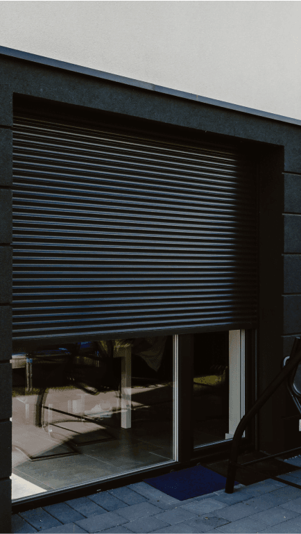 Nowoczesny budynek z dużymi oknami, zasłoniętymi aluminiowymi roletami zewnętrznymi w kolorze grafitowym.