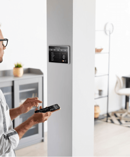 Mężczyzna stojący przy ścianie, obsługujący system inteligentnego domu za pomocą panelu dotykowego i smartfona.