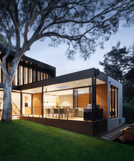 Nowoczesny dom z dużymi przeszkleniami, tarasem i zielonym ogrodem, oświetlony wieczorem.