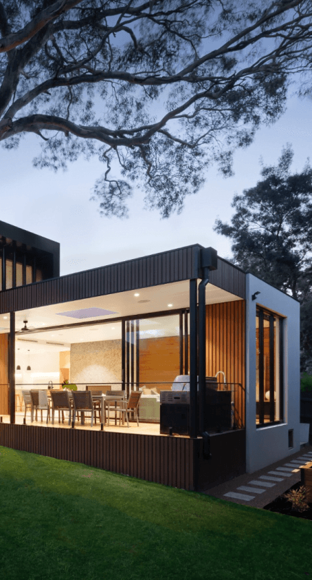 Nowoczesny dom z dużymi przeszkleniami, tarasem i zielonym ogrodem, oświetlony wieczorem.