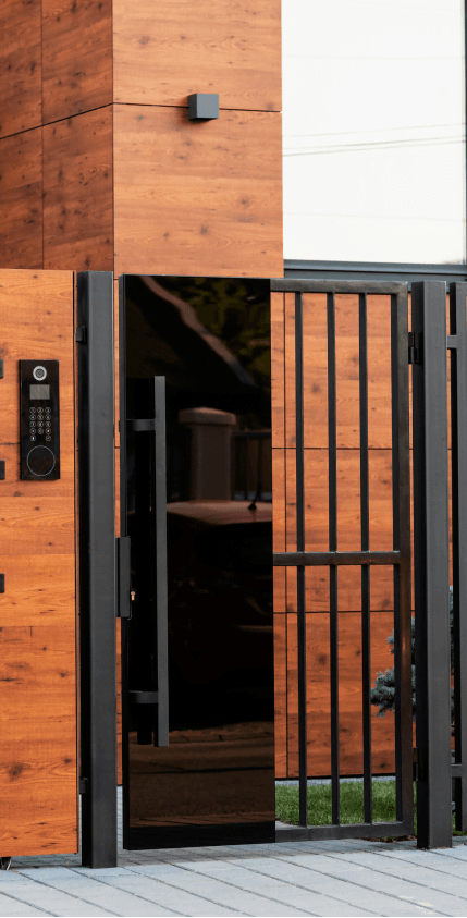 Drewniana elewacja budynku z zamkniętą, czarną furtką w stalowej ramie oraz panelem domofonowym z klawiaturą.