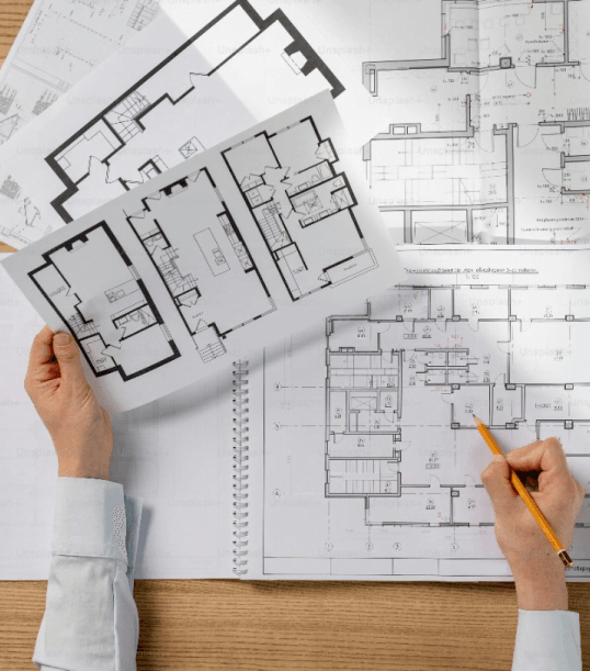 Osoba trzymająca i przeglądająca papierowe plany architektoniczne oraz rysująca ołówkiem po arkuszu na biurku.