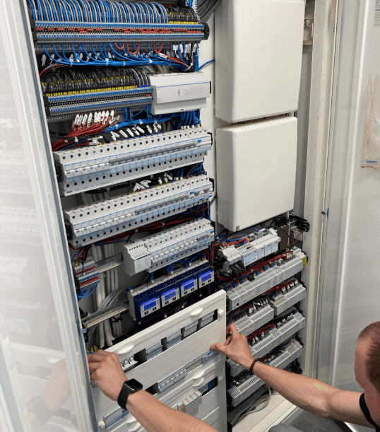 Osoba pracująca przy rozdzielnicy elektrycznej, w której znajduje się wiele kabli i komponentów instalacji inteligentnego domu.