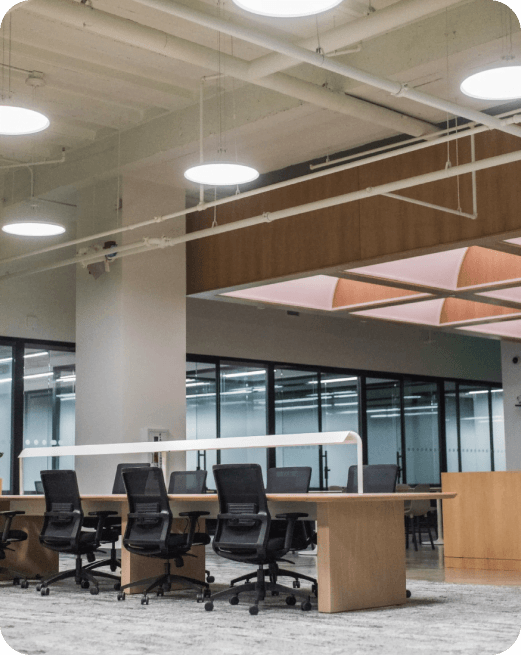 Nowoczesne biuro open space z drewnianymi stołami, czarnymi krzesłami biurowymi i industrialnym sufitem z odsłoniętą instalacją.