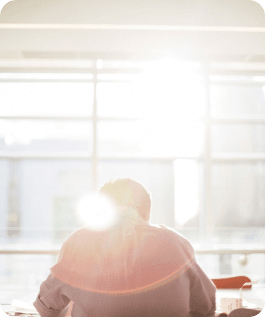 Sylwetka mężczyzny siedzącego przy biurku w jasnym pomieszczeniu, z intensywnym światłem słonecznym wpadającym przez okno.