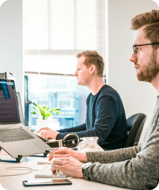 Dwóch mężczyzn pracujących przy komputerach w jasnym biurze, z rośliną na parapecie w tle.
