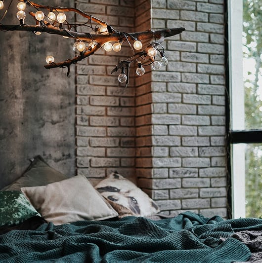 Łóżko z zielonym kocem, poduszkami oraz lampą z gałęzi i żarówek w stylu boho.